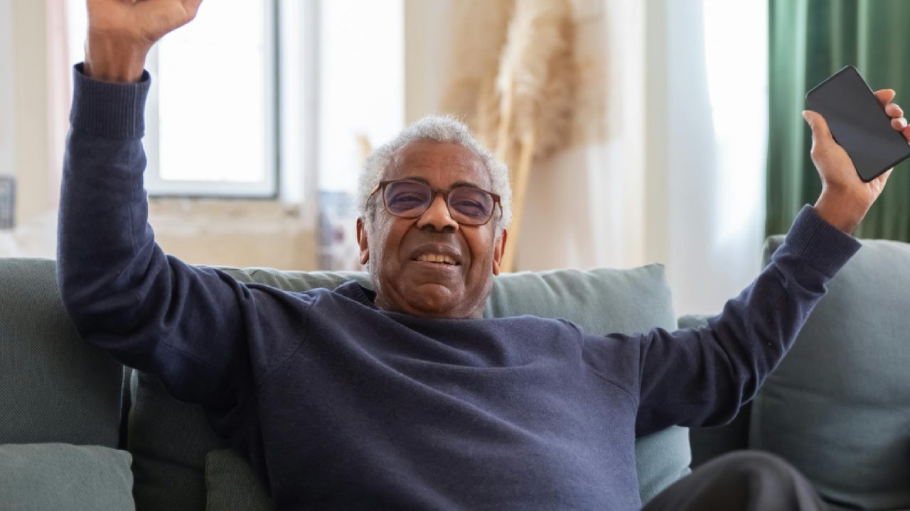 homem negro idoso de óculos e sueter azul comemorando ganho em jogo online com celular na mão